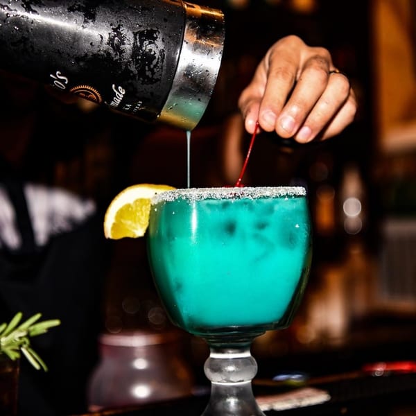 Bartender pouring a blue margarita from a shaker into a salt-rimmed glass with a lemon slice.