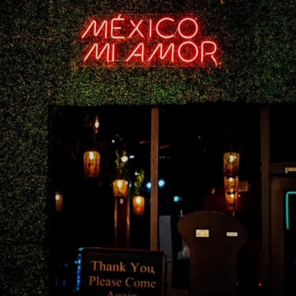 Red neon sign "MÉXICO MI AMOR" on a green hedge wall above a dark restaurant entrance.