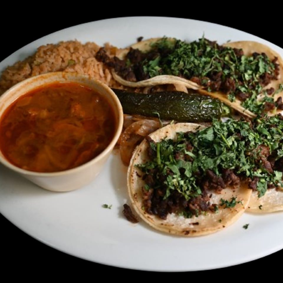 Three carne asada tacos with cilantro, rice, charro beans, and a grilled jalapeño on a white plate.