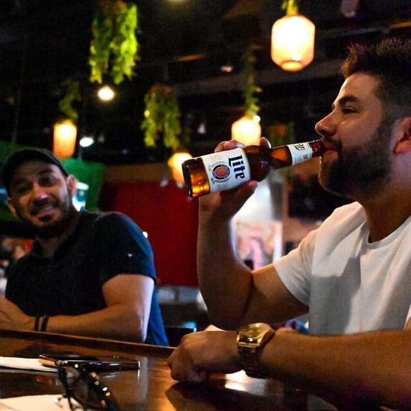 Man in white shirt sips a beer at a bar, another man smiles in the background.