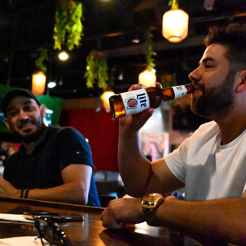 Man in white shirt sips a beer at a bar, another man smiles in the background.