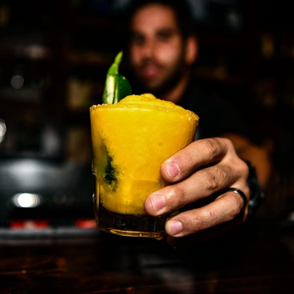 Bartender's hand holding a vibrant yellow frozen margarita garnished with a green chili.