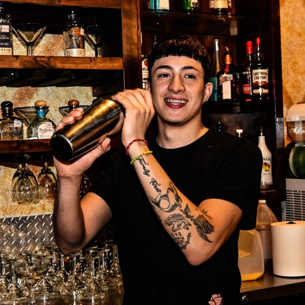 A smiling bartender with tattoos and braces shakes a cocktail shaker behind a bar with liquor bottles.
