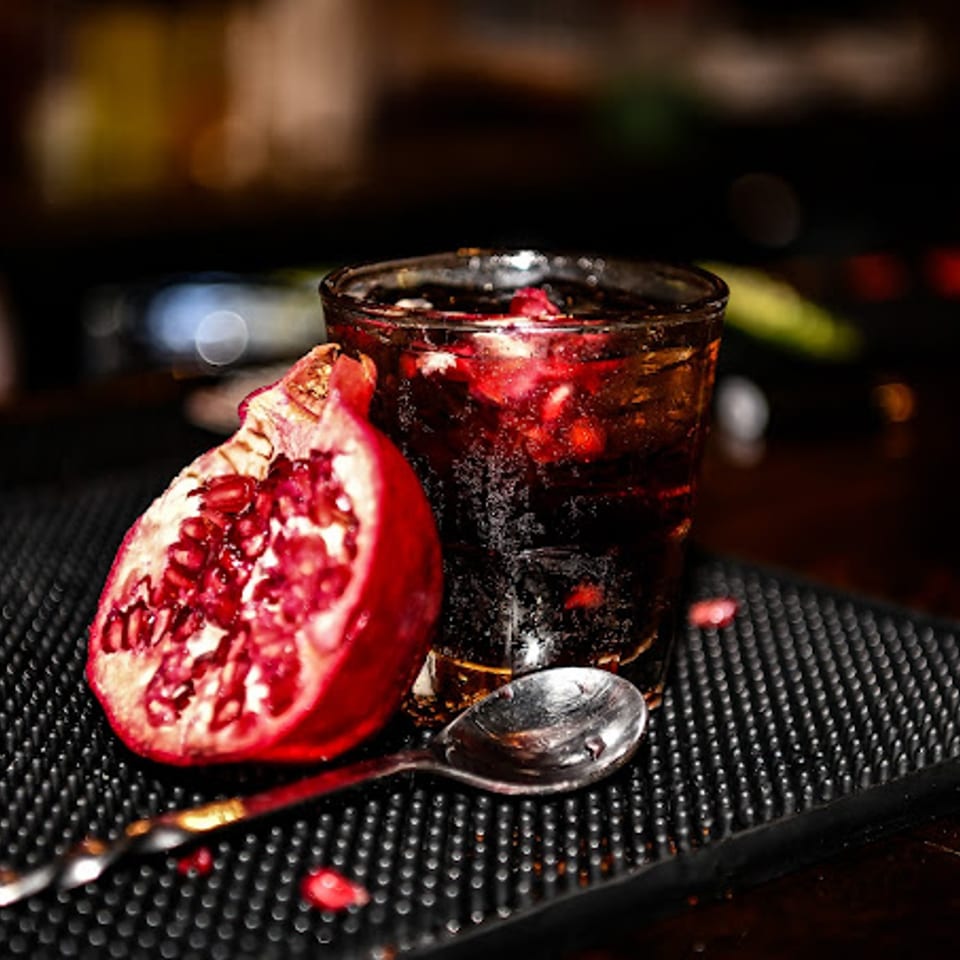 A dark red drink with pomegranate seeds, a cut pomegranate, and a spoon on a bar mat.
