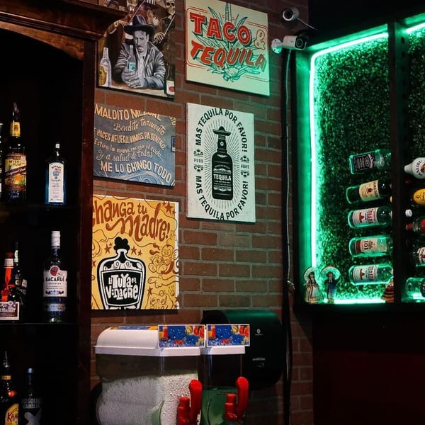 A bar wall with Mexican-themed art, liquor bottles, and a neon green lit display of bottles.